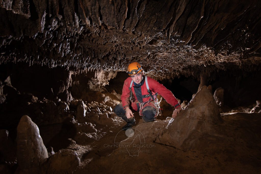 Spéléo en combinaison rouge accroupi dans un passage bas de la galerie. (Évent de Foussoubie, Ardèche)
