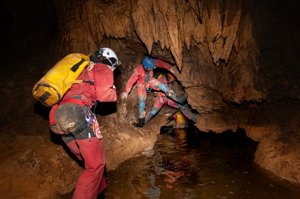 Passage bas lors de la traversée Cordier Évent inférieur. Des spéléo s’apprêtent à passer dans une vasque pleine d'eau. (Évent de Foussoubie, Ardèche)