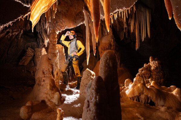 Un spéléo en jaune, entourée de spéléothèmes ocres. (Aven Grotte de l'Émeraude, Gard)