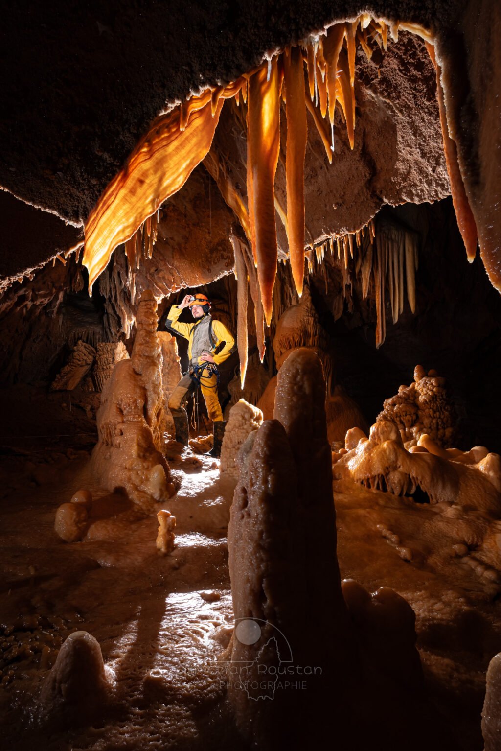Un spéléo en jaune, entourée de spéléothèmes ocres. (Aven Grotte de l'Émeraude, Gard)