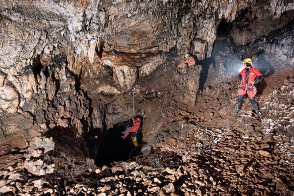 Deux spéléologues attendent sur la margelle du premier puits. Un troisième est sur la corde en train de descendre avec un kit jaune. (Aven Grotte Nouvelle, Ardèche)