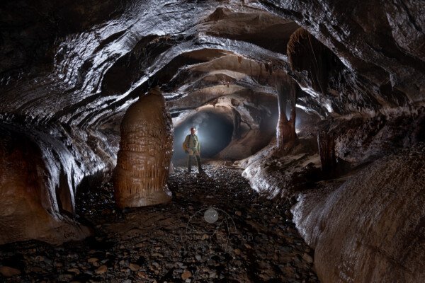 Une grosse stalagmite au bord de la galerie, spéléo en arrière plan dans la brume. (Réseau de la Cocalière, Résurgence de la Cotepatière, Aven de la Courcalhère, Ardèche)