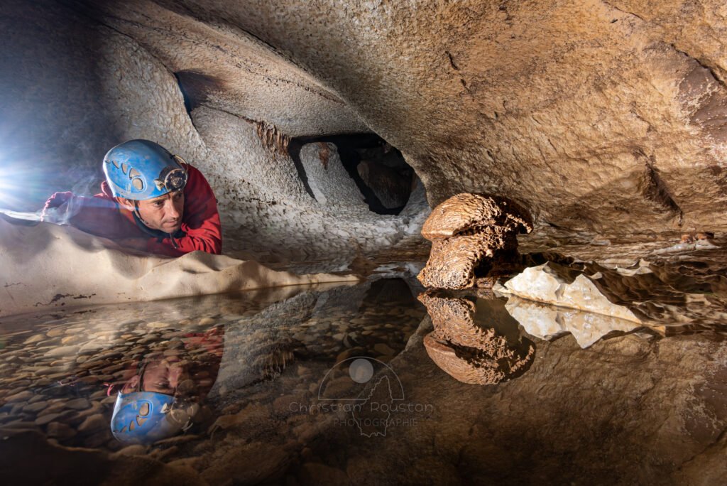 le spéléo regarde un champignon de pierre, le reflet de la scène est visible dans une marmite. (Évent de Peyrejal, Ardèche)