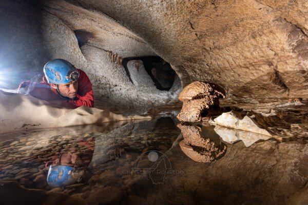 le spéléo regarde un champignon de pierre, le reflet de la scène est visible dans une marmite. (Évent de Peyrejal, Ardèche)