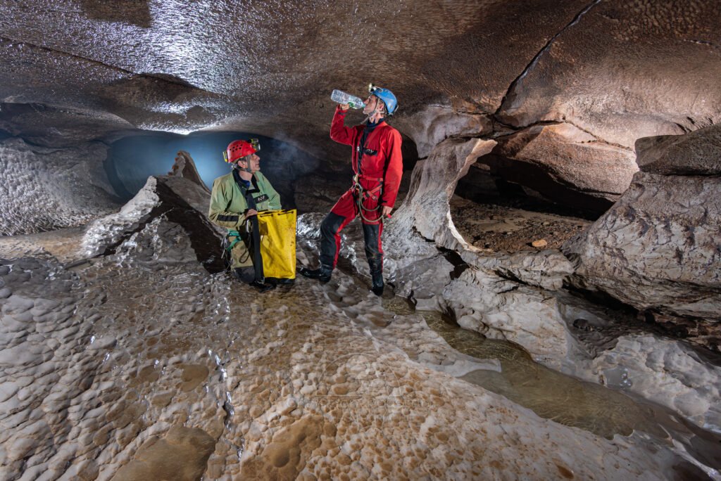 Spéléo avec sac ouvert, un autre boit de l'eau à la bouteille (Évent de Peyrejal, Ardèche)