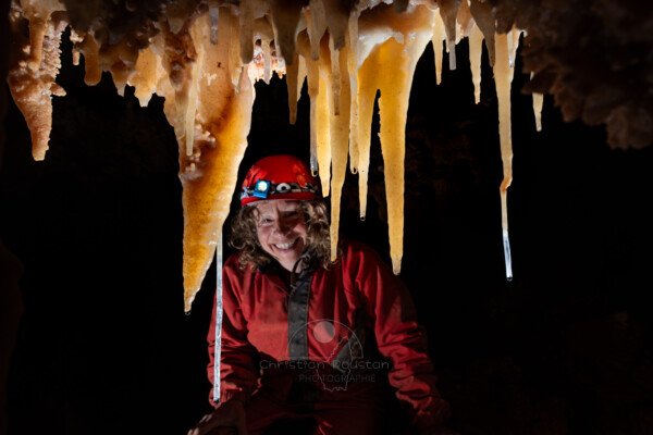 Spéléologue souriante derrière un rideau de concrétions (Spéléothème, Grotte du Philosophe, Gard)